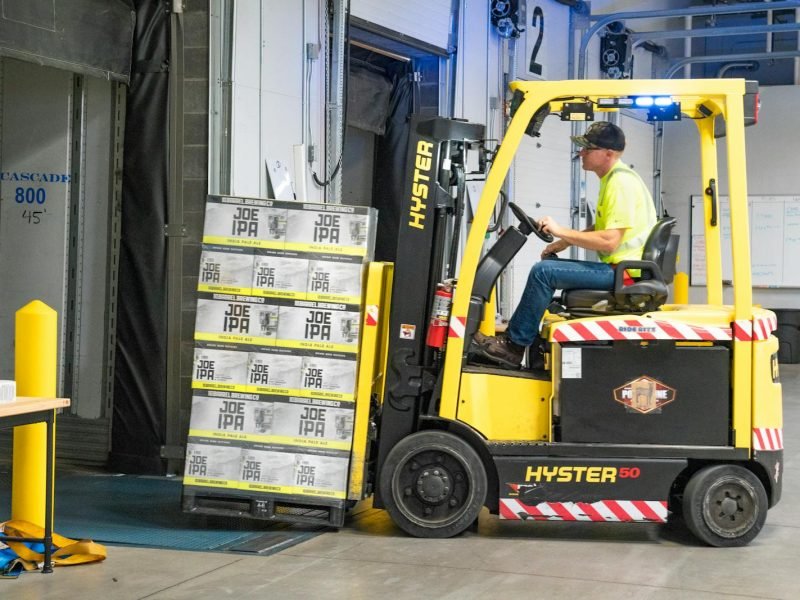 A worker drives a Hyster forklift moving Joe IPA boxes in a warehouse.
